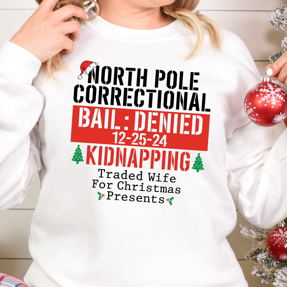 May include: White sweatshirt with a humorous Christmas-themed graphic. The text reads "NORTH POLE CORRECTIONAL BAIL: DENIED 12-25-24 KIDNAPPING Traded Wife For Christmas Presents" in black and red. A Santa hat and Christmas trees are also included.