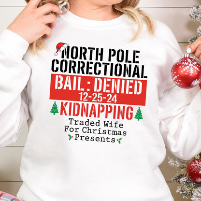 May include: White sweatshirt with a humorous Christmas-themed graphic. The text reads "NORTH POLE CORRECTIONAL BAIL: DENIED 12-25-24 KIDNAPPING Traded Wife For Christmas Presents" in black and red. A Santa hat and Christmas trees are also included.