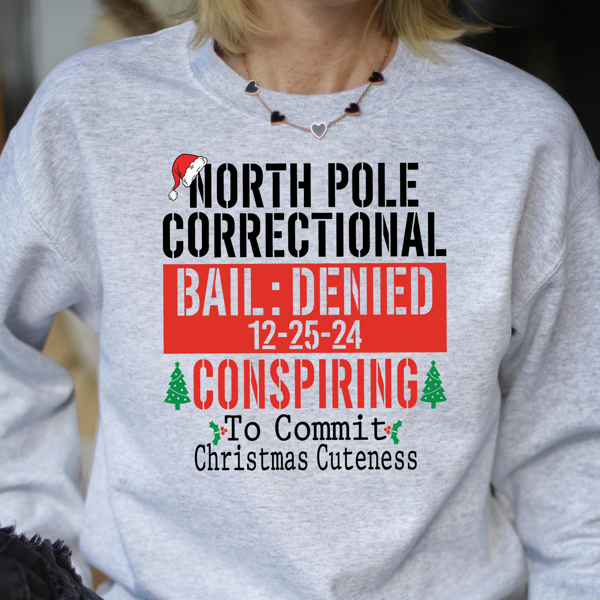 May include: A light gray sweatshirt with the text "NORTH POLE CORRECTIONAL BAIL: DENIED 12-25-24 CONSPIRING To Commit Christmas Cuteness" in black and red. A Santa hat graphic is above the text.