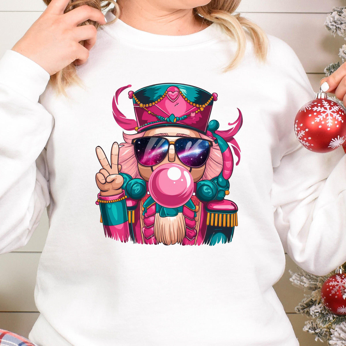 May include: White sweatshirt featuring a colorful illustration of a nutcracker wearing sunglasses and blowing a bubblegum bubble. The nutcracker is in a peace sign pose. The design includes pink, teal, and gold accents, perfect for the holiday season.