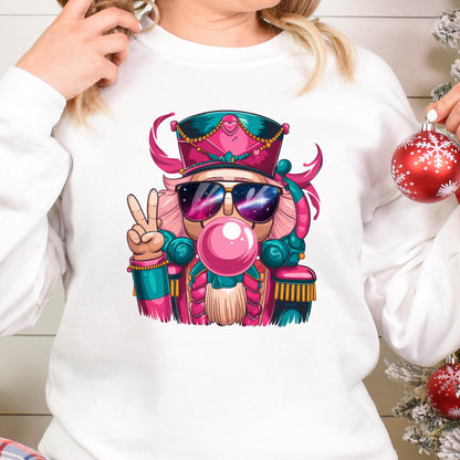 May include: White sweatshirt featuring a colorful illustration of a nutcracker wearing sunglasses and blowing a bubblegum bubble. The nutcracker is in a peace sign pose. The design includes pink, teal, and gold accents, perfect for the holiday season.