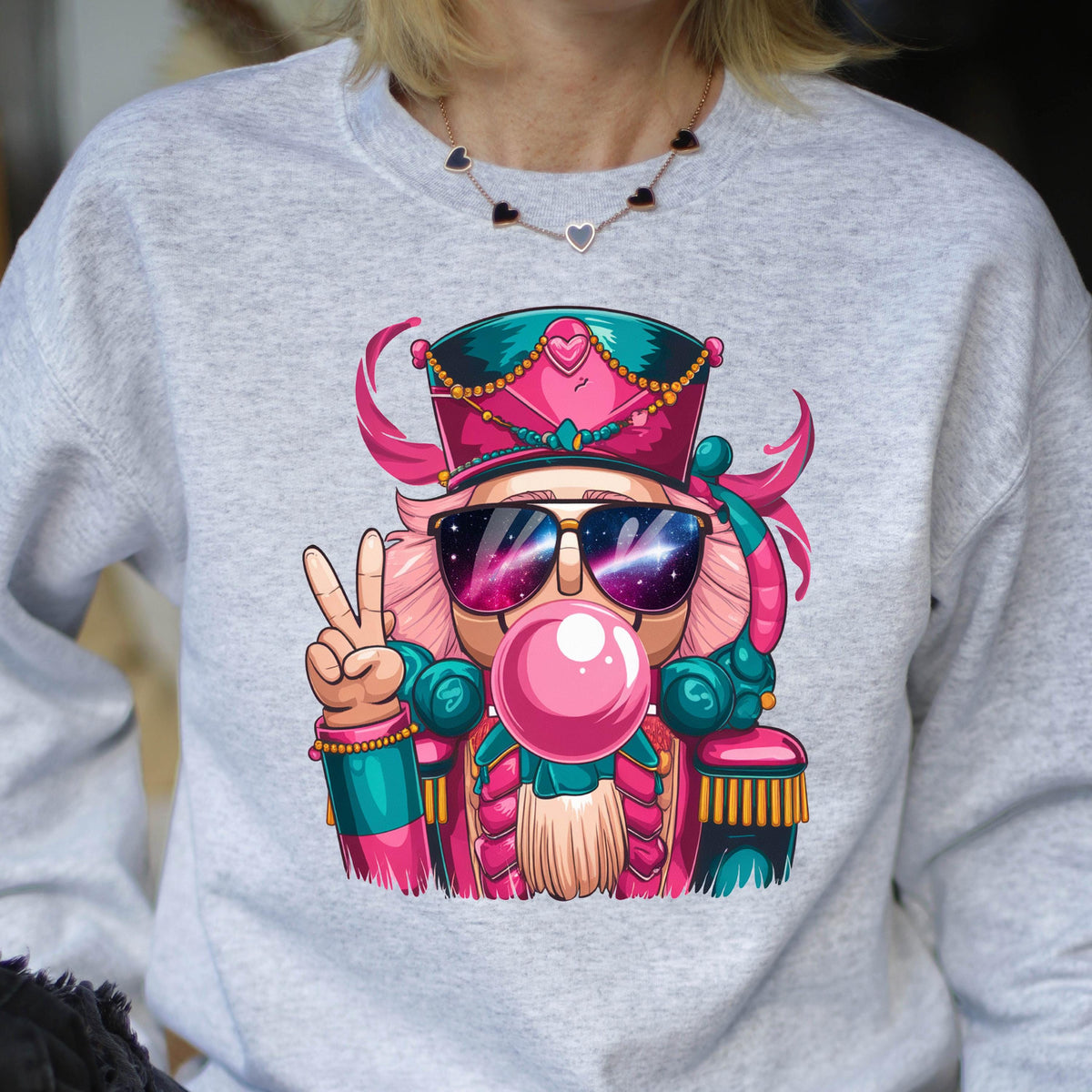 May include: A light gray sweatshirt featuring a colorful cartoon nutcracker wearing sunglasses with a galaxy design, blowing a bubblegum bubble, and giving a peace sign. The nutcracker is wearing a pink and teal uniform and hat.