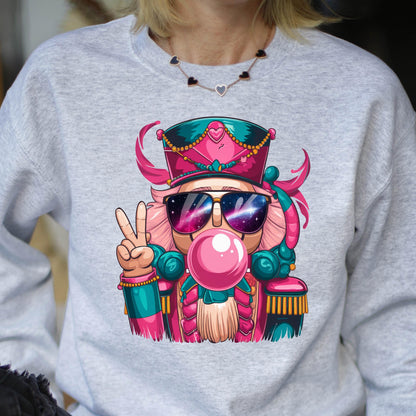 May include: A light gray sweatshirt featuring a colorful cartoon nutcracker wearing sunglasses with a galaxy design, blowing a bubblegum bubble, and giving a peace sign. The nutcracker is wearing a pink and teal uniform and hat.