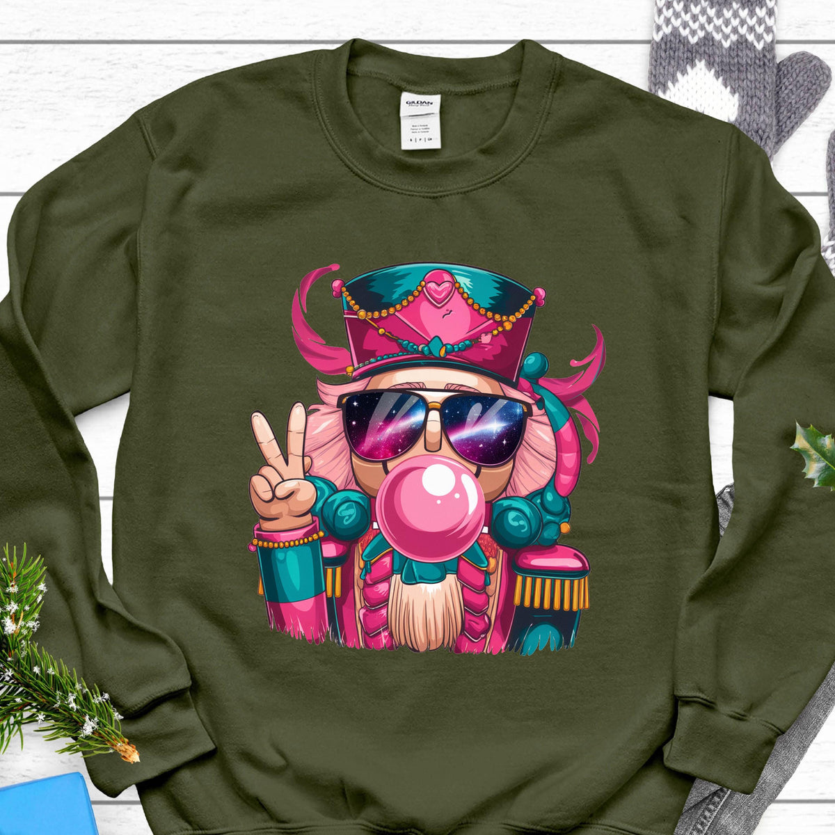 May include: An olive green sweatshirt featuring a cartoon nutcracker wearing sunglasses and blowing a bubblegum bubble. The nutcracker is wearing a pink and teal hat and uniform, and is giving a peace sign with one hand.