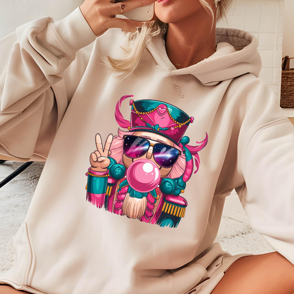 May include: Beige hoodie featuring a colorful graphic of a nutcracker wearing sunglasses and blowing a bubble. The nutcracker is depicted with pink hair, a teal and pink hat, and a peace sign. The hoodie has a drawstring hood and a kangaroo pocket.