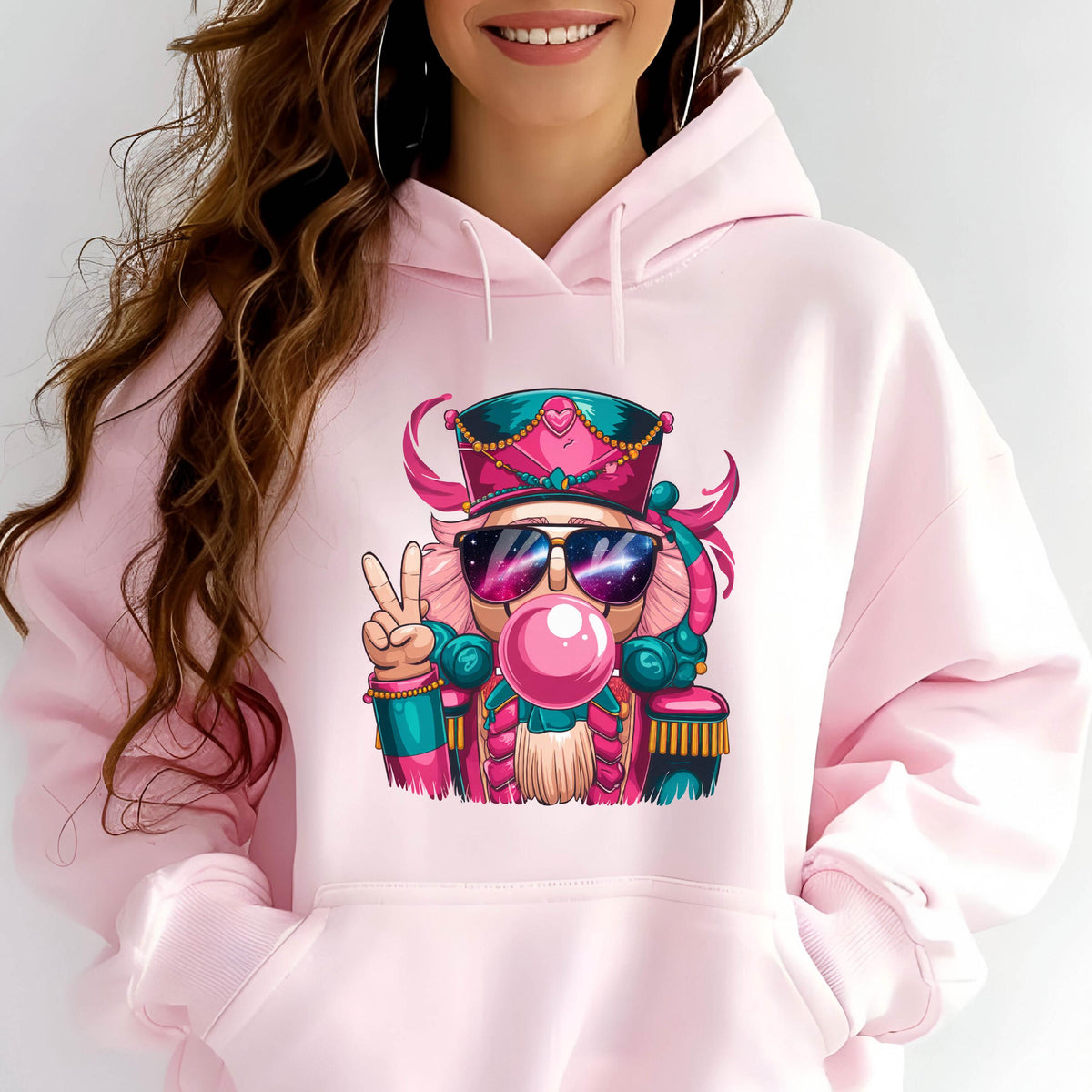 May include: A light pink hoodie featuring a colorful cartoon nutcracker with sunglasses blowing a bubblegum bubble. The nutcracker is wearing a pink and teal hat and uniform, and is giving a peace sign.