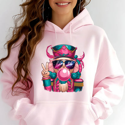 May include: A light pink hoodie featuring a colorful cartoon nutcracker with sunglasses blowing a bubblegum bubble. The nutcracker is wearing a pink and teal hat and uniform, and is giving a peace sign.