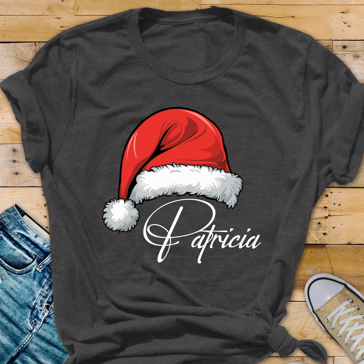 May include: A dark gray short-sleeved t-shirt featuring a graphic of a red Santa hat with white fur trim and a pom-pom. Below the hat, the name Patricia is printed in a white cursive script font across the center of the chest area.