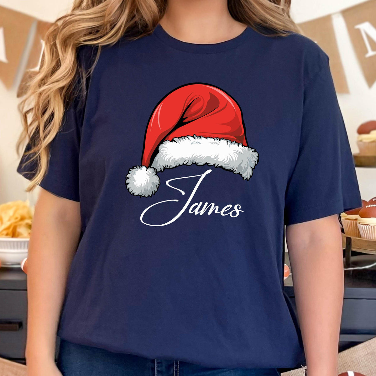 May include: A navy blue short-sleeve t-shirt featuring a graphic of a red Santa hat with a white fur trim and pom-pom. Below the hat, the name James is printed in a white cursive script font. The design is centered on the front of the shirt.