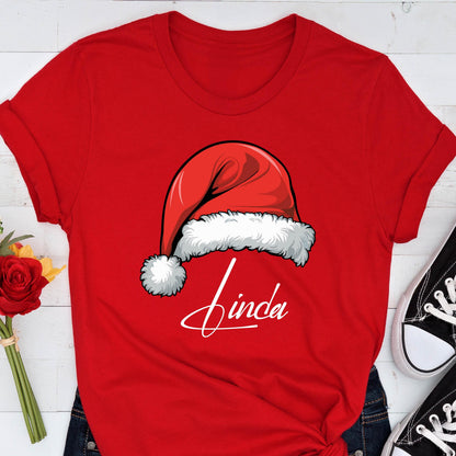 May include: A bright red short-sleeved t-shirt featuring a graphic of a red Santa hat with a white fur trim and pom-pom. Below the hat, the name Linda is printed in a white cursive script font. The shirt has a crew neckline and cuffed sleeves.