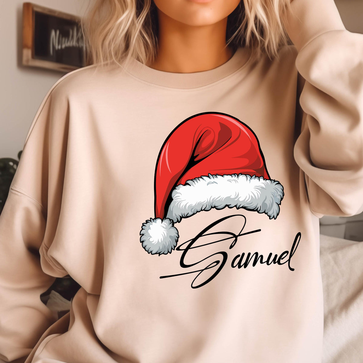 May include: A beige crewneck sweatshirt featuring a graphic of a red Santa hat with a white fur trim and pom-pom. Below the hat, the name Samuel is printed in a black cursive script font. The sweatshirt has long sleeves and a ribbed neckline.