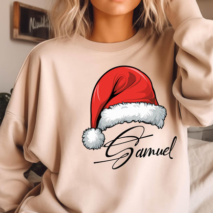 May include: A beige crewneck sweatshirt featuring a graphic of a red Santa hat with a white fur trim and pom-pom. Below the hat, the name Samuel is printed in a black cursive script font. The sweatshirt has long sleeves and a ribbed neckline.