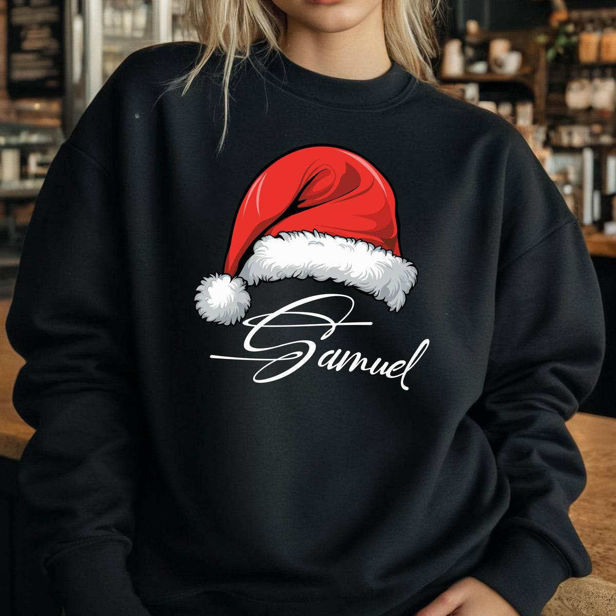 May include: A black long-sleeve sweatshirt featuring a graphic of a red Santa hat with a white fur trim and pom-pom. Below the hat, the name Samuel is printed in a white cursive script font. The garment has a crew neckline and ribbed cuffs.