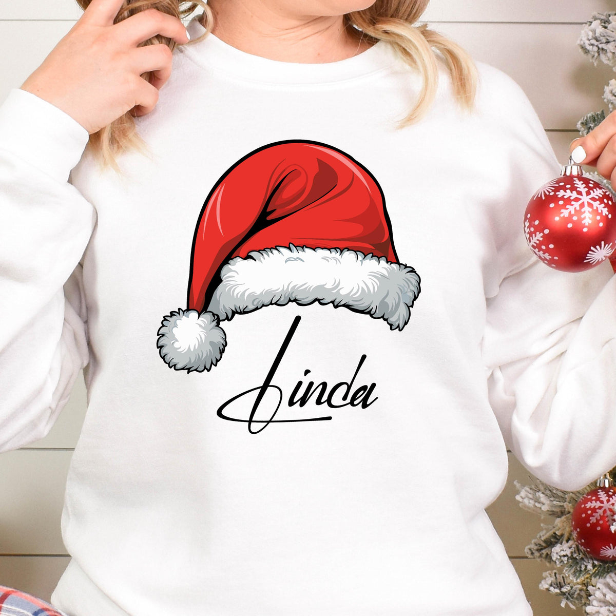 May include: A white long-sleeve sweatshirt featuring a graphic of a red Santa hat with a white fur trim and pom-pom. Below the hat, the name Linda is printed in a black cursive script font. The design is centered on the front of the garment.