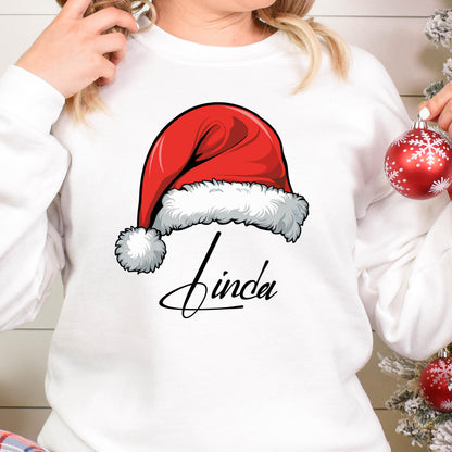 May include: A white long-sleeve sweatshirt featuring a graphic of a red Santa hat with a white fur trim and pom-pom. Below the hat, the name Linda is printed in a black cursive script font. The design is centered on the front of the garment.