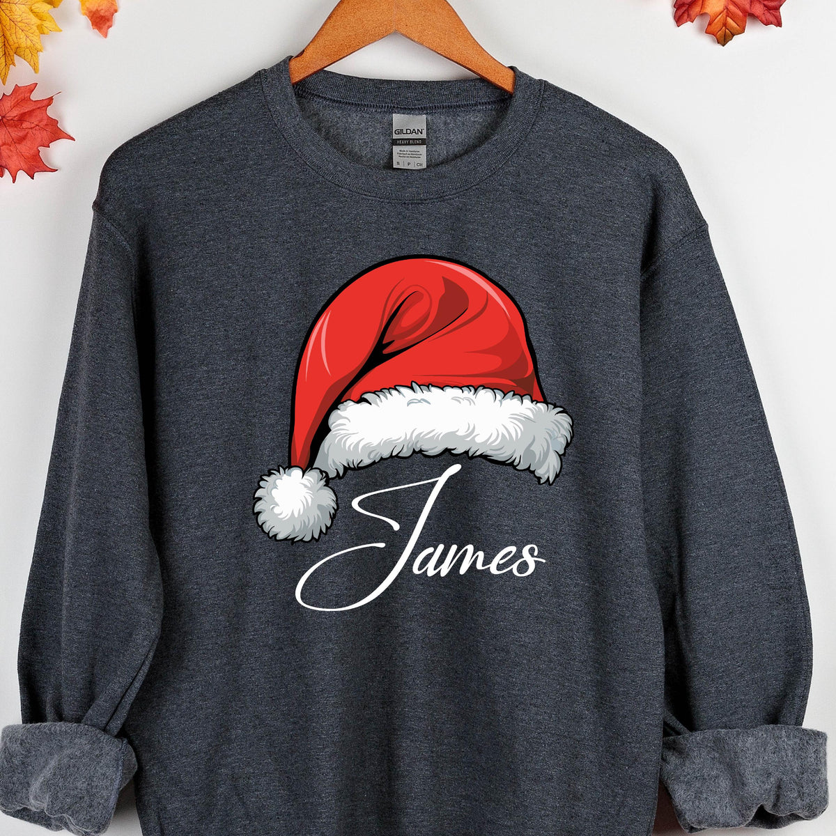 May include: A dark heather gray crewneck sweatshirt featuring a graphic of a red Santa hat with white fur trim. Below the hat, the name James is printed in a white cursive script font. The garment has long sleeves and a ribbed neckline.