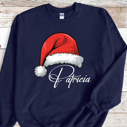 May include: A navy blue crewneck sweatshirt featuring a graphic of a red Santa hat with white fur trim and a pom-pom. Below the hat, the name Patricia is printed in a white cursive script font. The garment has long sleeves and a ribbed collar.
