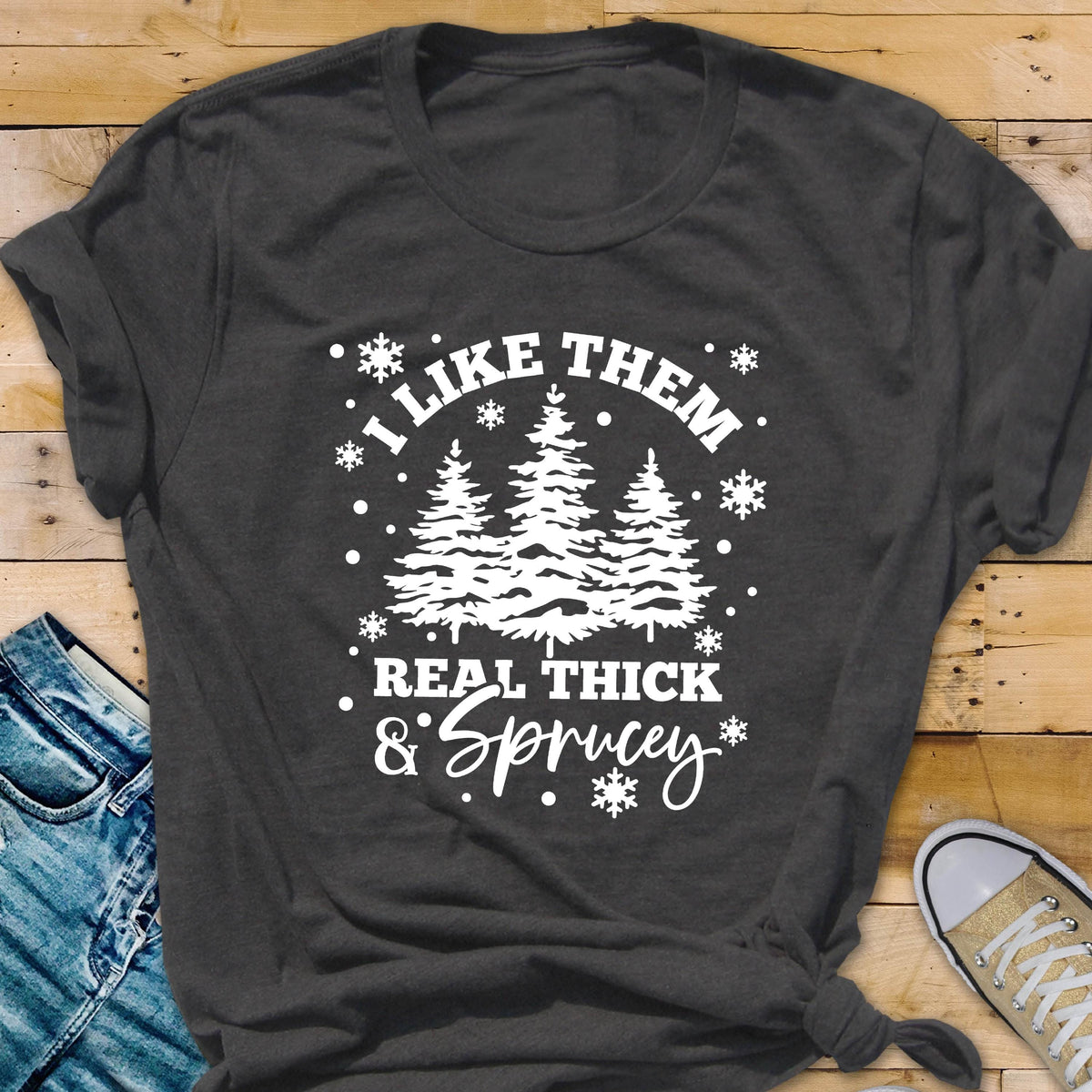 May include: Dark gray t-shirt with a white graphic featuring evergreen trees and the text "I LIKE THEM REAL THICK & SPRUCEY" in a whimsical font. The design includes snowflakes. The shirt is folded.