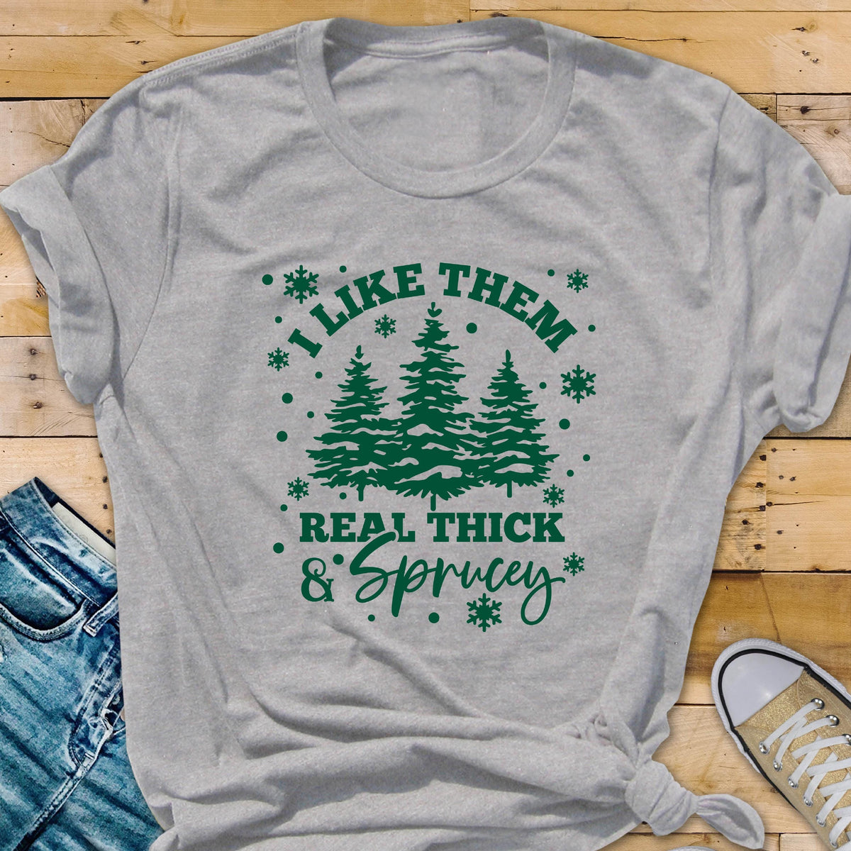 May include: A heather gray t-shirt with a green graphic that reads "I LIKE THEM REAL THICK & SPRUCEY" with illustrations of pine trees and snowflakes. The shirt is folded and tied at the bottom.