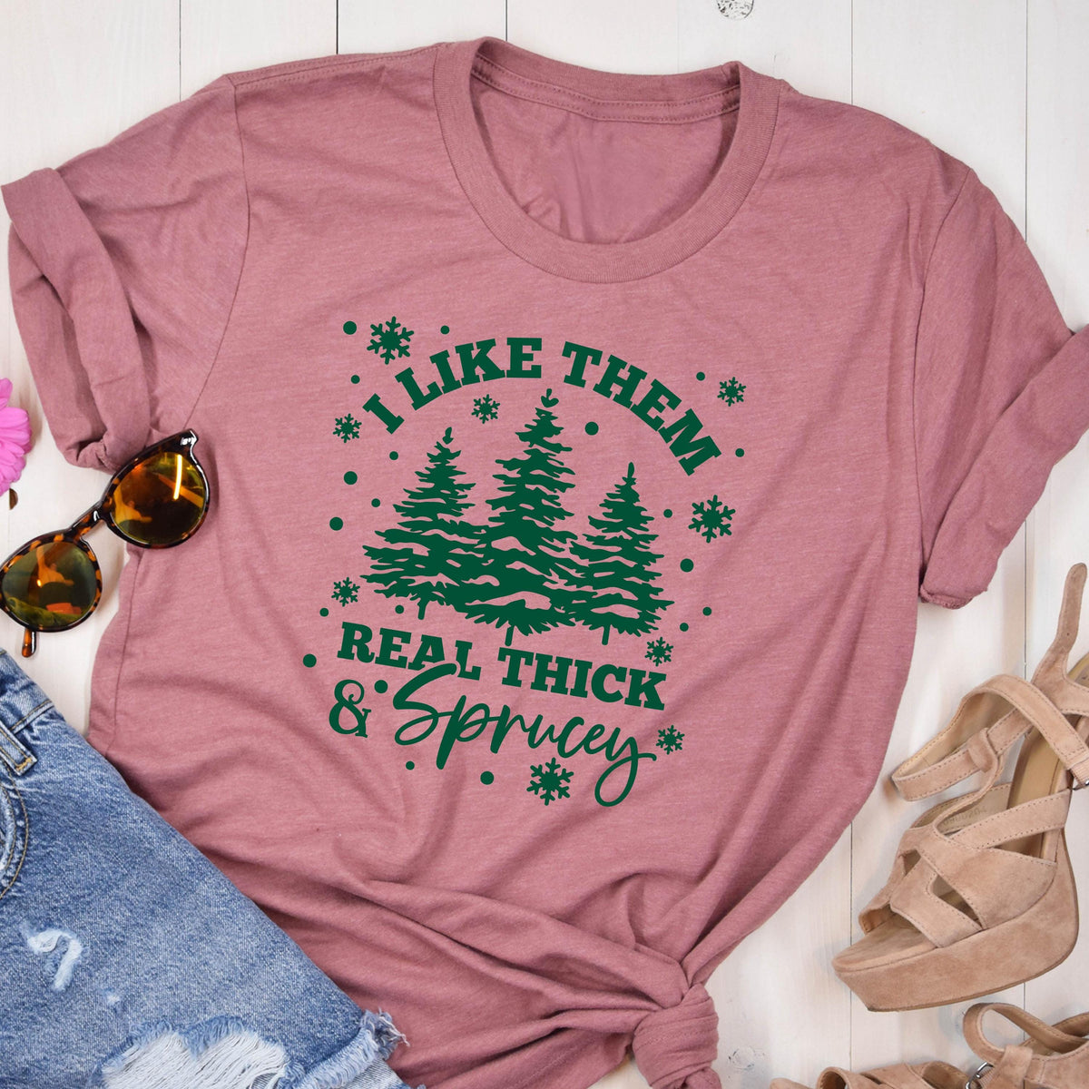 May include: A dusty rose t-shirt with the phrase "I LIKE THEM REAL THICK & Sprucey" in green. The design includes three green pine trees and snowflake accents. The shirt is folded and styled with sunglasses and denim shorts.