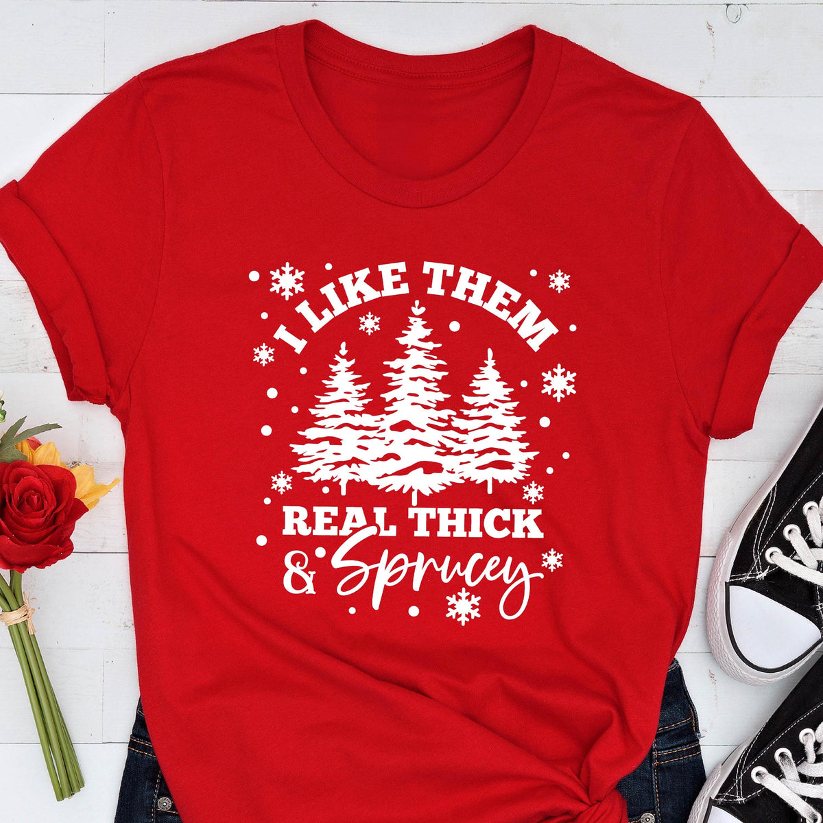 May include: A red t-shirt with a white graphic that reads "I LIKE THEM REAL THICK & Sprucey" with images of pine trees and snowflakes. The shirt is folded at the sleeves.