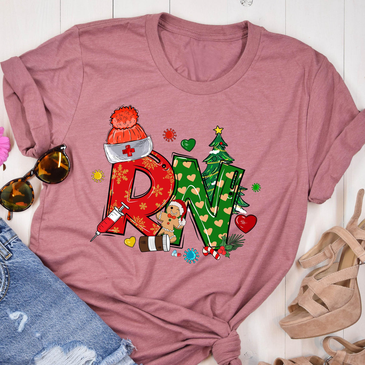 May include: A dusty rose t-shirt featuring a festive design for a Registered Nurse. The design includes the letters "RN" in red and green, decorated with snowflakes, hearts, a Christmas tree, and a gingerbread man. A syringe and a nurse's hat are also included.
