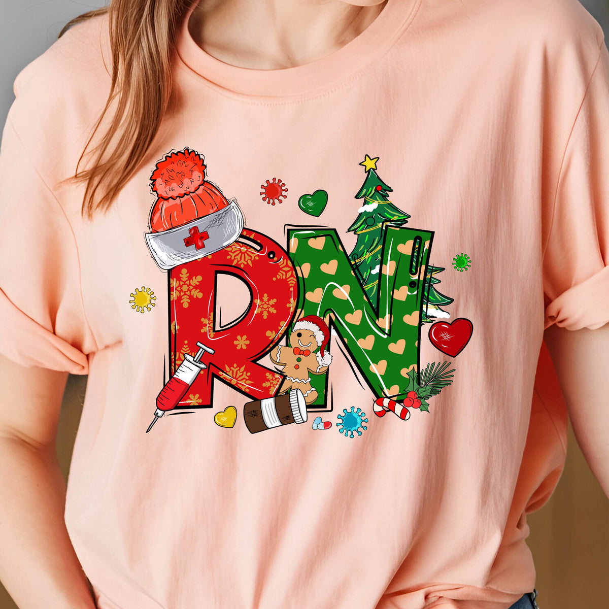 May include: A light peach-colored t-shirt featuring a festive design. The graphic spells out "RN" with a red "D" adorned with snowflakes and a nurse's hat. The green "N" is decorated with hearts and a Christmas tree. A gingerbread man and medical elements complete the design.