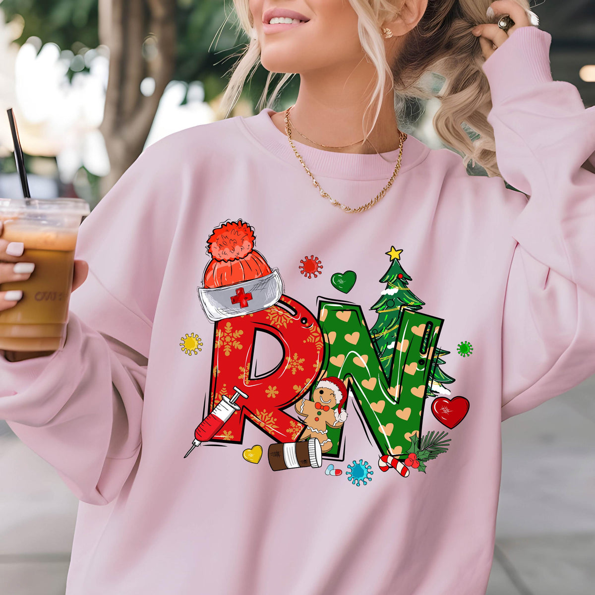 May include: A light pink crewneck sweatshirt featuring a graphic print of the letters RN. The R is red with snowflake patterns and a syringe, while the N is green with heart patterns and a Christmas tree. Includes a nurse hat and gingerbread man.
