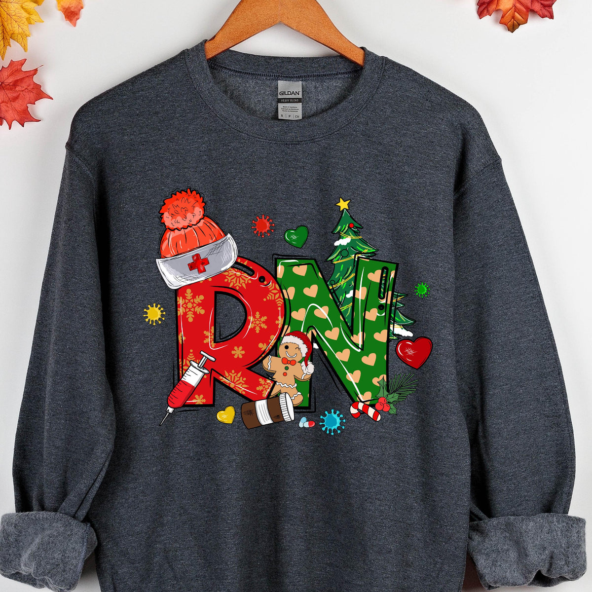 May include: A dark gray crewneck sweatshirt featuring a large graphic with the letters RN. The R is red with snowflake patterns and a nurse cap, while the N is green with heart patterns and a Christmas tree. Includes a gingerbread man and syringe.