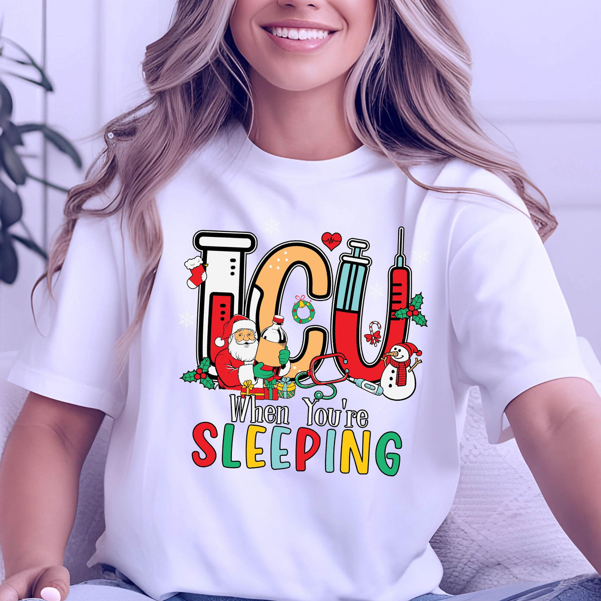May include: White t-shirt with a colorful graphic design featuring the letters "ICU" in a medical theme, with Santa Claus, a snowman, and the text "When You're Sleeping". The design incorporates medical equipment.