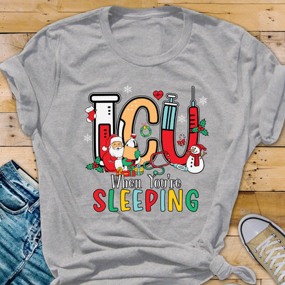 May include: A gray t-shirt with a colorful graphic that reads "ICU When You're Sleeping." The design includes medical equipment, snowflakes, and Christmas-themed elements like Santa Claus and a snowman.