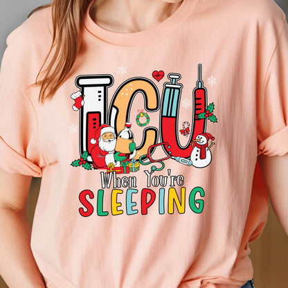 May include: A light pink t-shirt featuring a colorful graphic with the letters "ICU" and the phrase "When You're Sleeping." The design includes medical equipment, Santa Claus, a snowman, and snowflakes, creating a festive, medical theme.