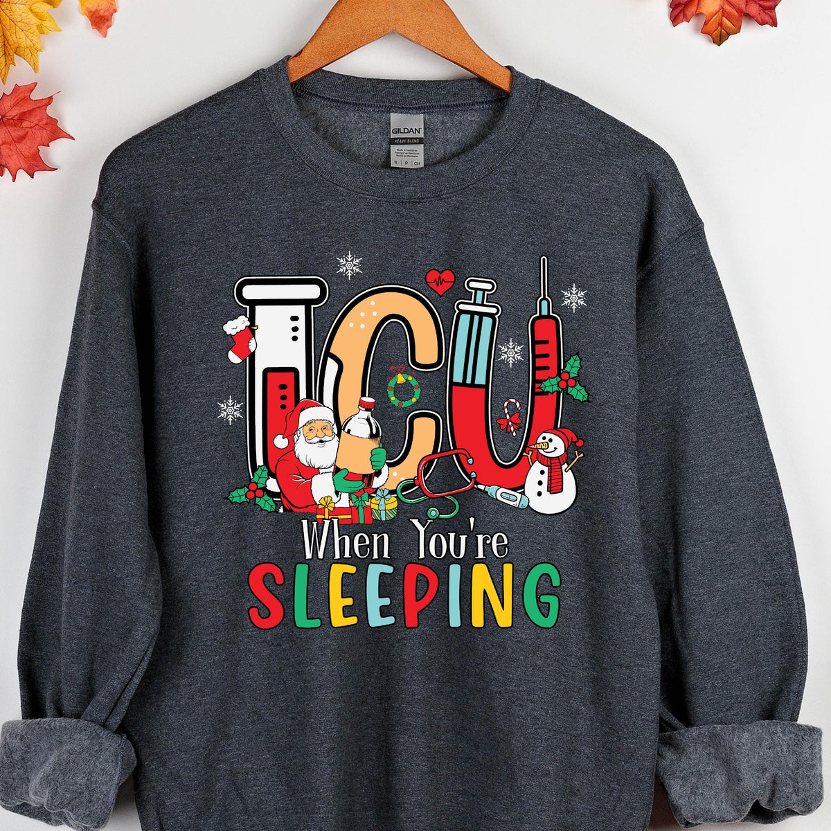 May include: Dark gray sweatshirt with a colorful graphic. The graphic features the letters "ICU" in orange and red, along with Santa Claus, a snowman, and medical equipment. The text "When You're SLEEPING" is below the graphic.