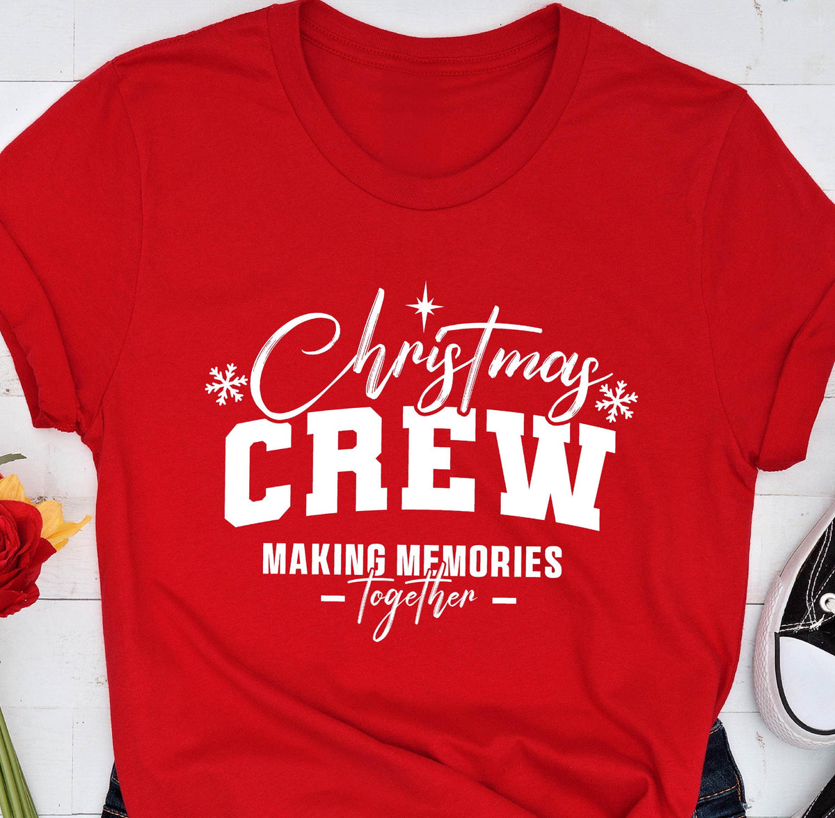 May include: A bright red short-sleeved t-shirt featuring white screen-printed text that reads Christmas CREW MAKING MEMORIES Together, accented with two white snowflake icons and a small star symbol centered above the word Christmas.