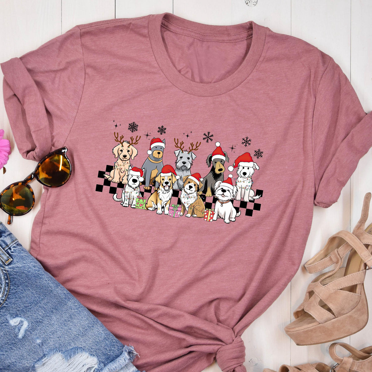 May include: A dusty rose t-shirt featuring a graphic of various dog breeds wearing Santa hats and reindeer antlers. The dogs are arranged on a black and white checkered pattern, with snowflakes above them. The shirt is a crew neck style.
