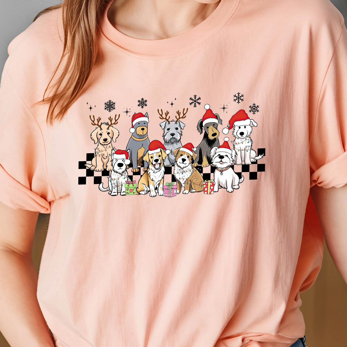 May include: A light peach-colored t-shirt featuring a festive graphic of various dog breeds wearing Santa hats and reindeer antlers. The dogs are arranged in a row on a black and white checkered pattern, with snowflakes above.