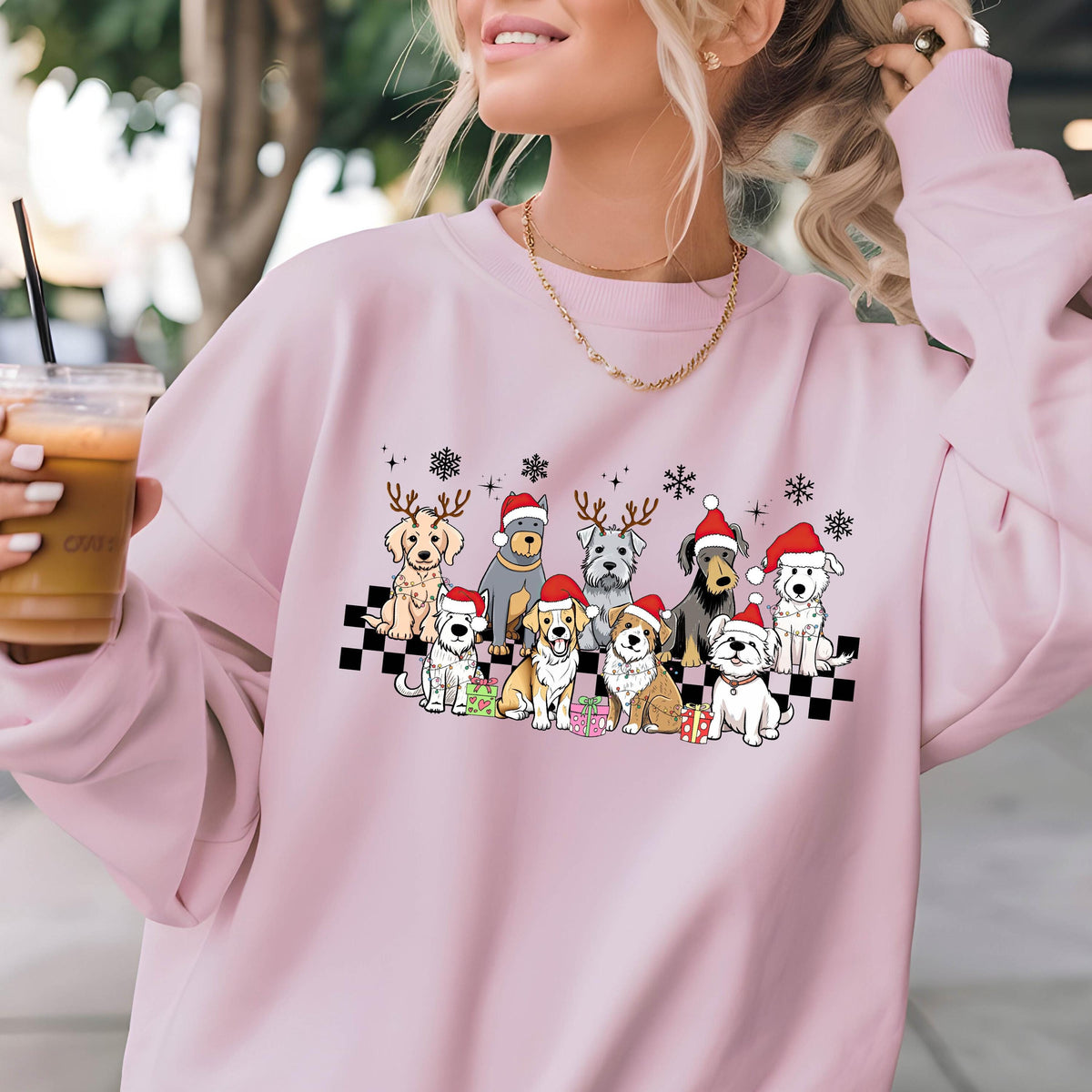 May include: A light pink sweatshirt featuring a festive graphic of various dog breeds wearing Santa hats and reindeer antlers. The dogs are arranged on a black and white checkered pattern, with snowflakes above them. A gold chain is visible.