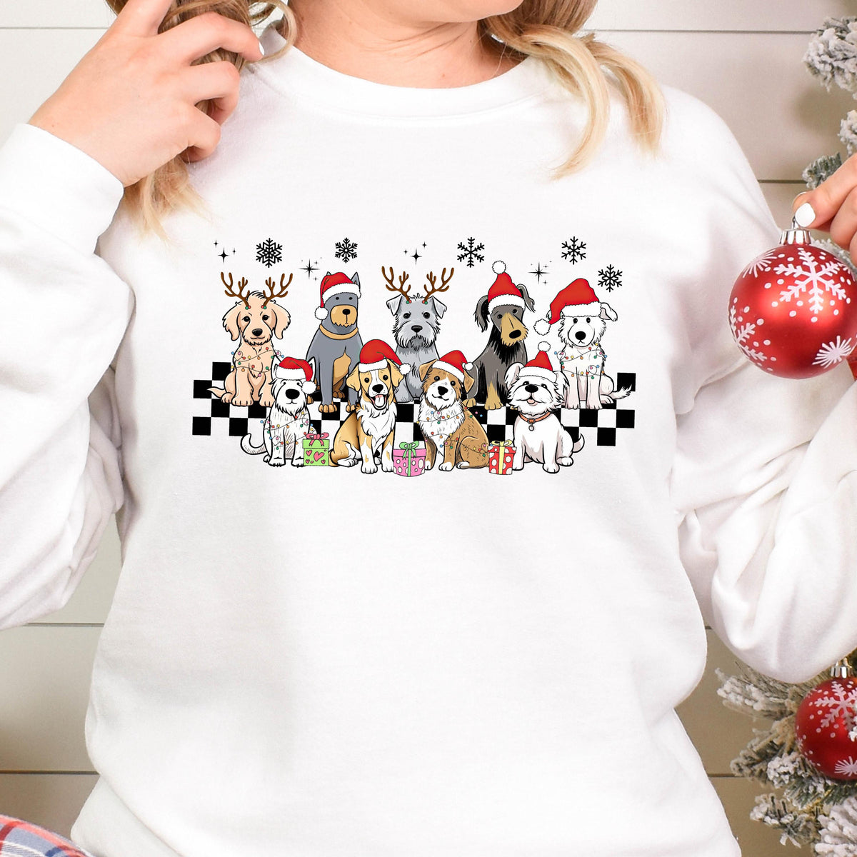 May include: White sweatshirt featuring a festive design of various dog breeds wearing Santa hats and reindeer antlers. The dogs are arranged on a black and white checkered pattern, with snowflakes and wrapped gifts. A cheerful holiday theme.