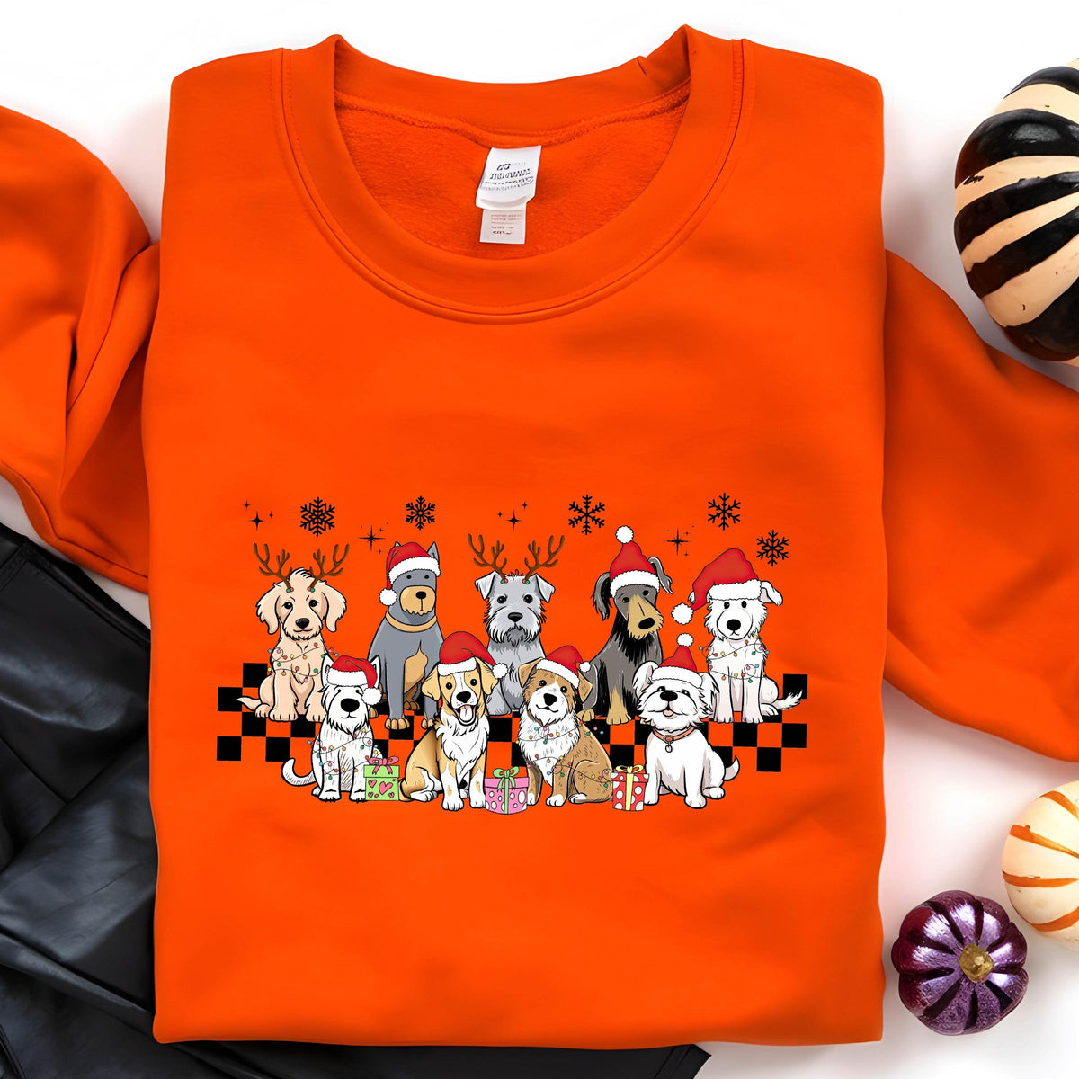 May include: An orange sweatshirt featuring a festive design of various dog breeds wearing Santa hats and reindeer antlers. The dogs are arranged on a black and white checkered pattern, with snowflakes and wrapped gifts. A cozy and cheerful holiday theme.