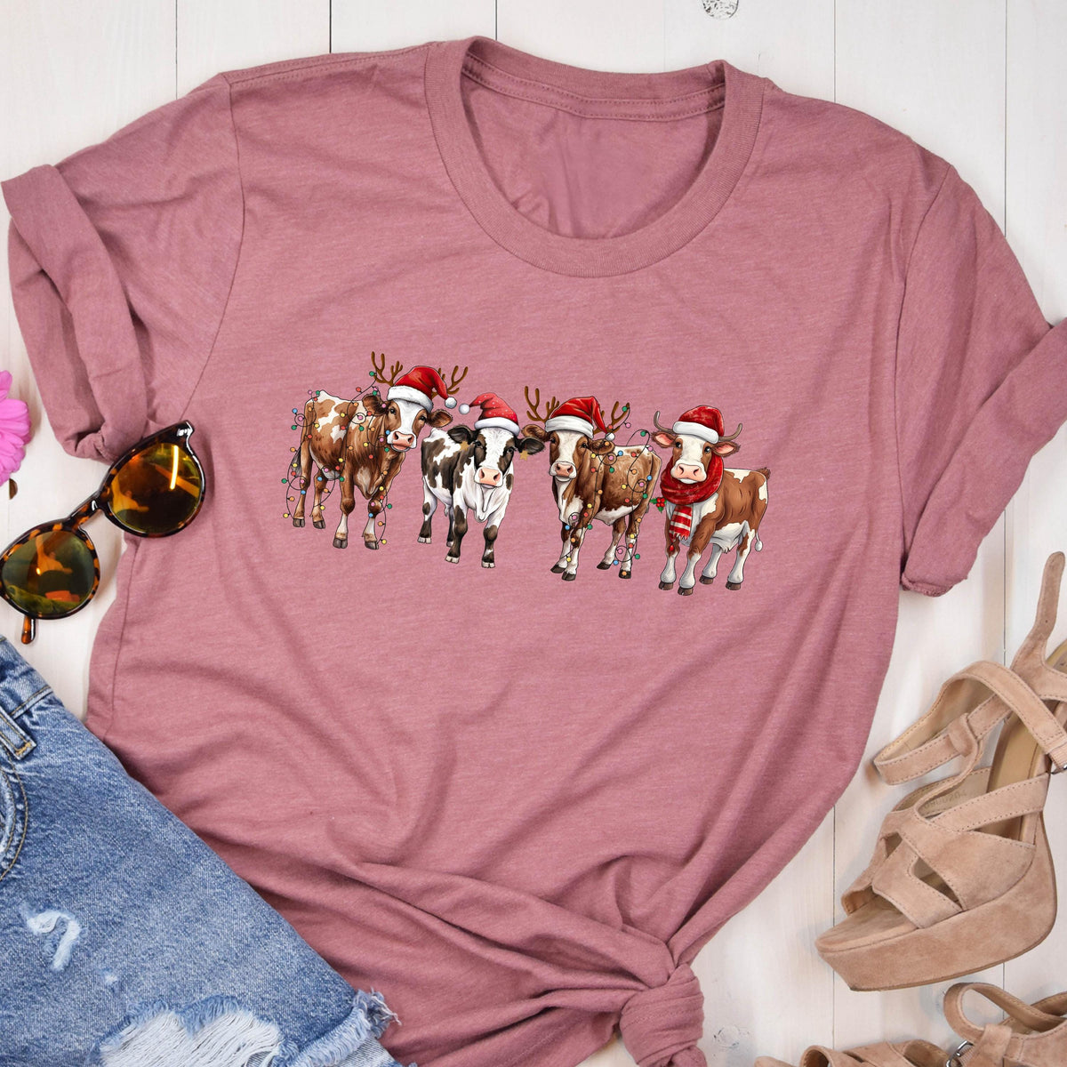 May include: A mauve-colored t-shirt featuring a festive graphic of six cows adorned with Santa hats, reindeer antlers, and Christmas lights. One cow wears a red and white striped scarf. The cows are in a row, creating a holiday theme.