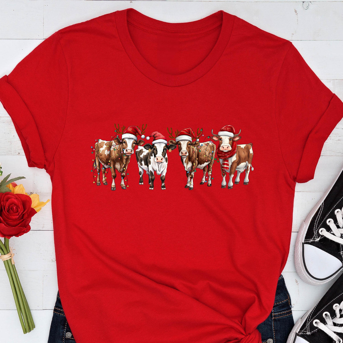 May include: A red t-shirt featuring a festive design of five cows. Each cow is adorned with a Santa hat, and some have reindeer antlers or scarves. The cows are in a row, creating a whimsical holiday theme.