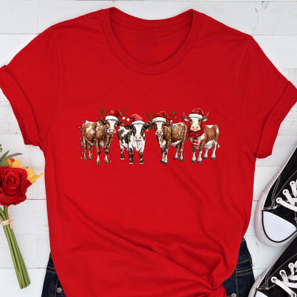 May include: A red t-shirt featuring a festive design of five cows. Each cow is adorned with a Santa hat, and some have reindeer antlers or scarves. The cows are in a row, creating a whimsical holiday theme.