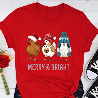 May include: Red t-shirt featuring three cartoon chickens wearing winter hats. The chickens are brown, white, and white with a blue hat. The text "MERRY & BRIGHT" is printed below the chickens in a silver color.