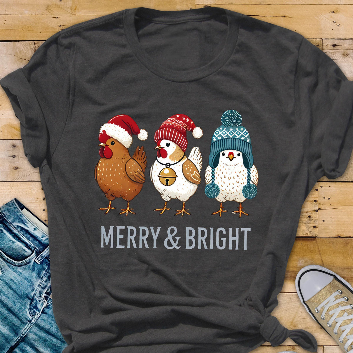 May include: Dark gray t-shirt featuring three cartoon chickens wearing festive winter hats. The chickens are brown, white, and blue, with a red Santa hat, a red and white knit cap, and a blue knit cap. The text "MERRY & BRIGHT" is printed below the chickens.