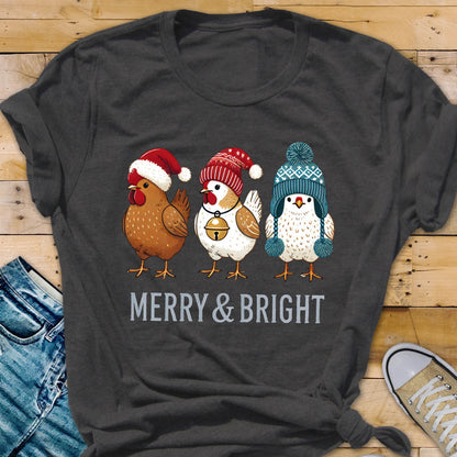 May include: Dark gray t-shirt featuring three cartoon chickens wearing festive winter hats. The chickens are brown, white, and blue, with a red Santa hat, a red and white knit cap, and a blue knit cap. The text "MERRY & BRIGHT" is printed below the chickens.