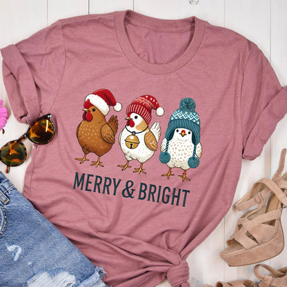 May include: A dusty rose t-shirt featuring a graphic of three chickens wearing festive winter hats. The chickens are brown, white, and blue, with the text "MERRY & BRIGHT" printed below in black.