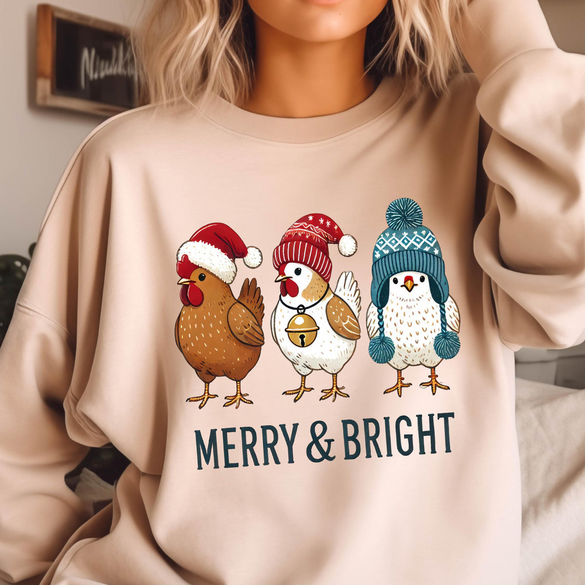 May include: A beige sweatshirt with a Christmas-themed design featuring three chickens wearing Santa hats and a blue and white winter hat. The text "Merry & Bright" is printed below the chickens.
