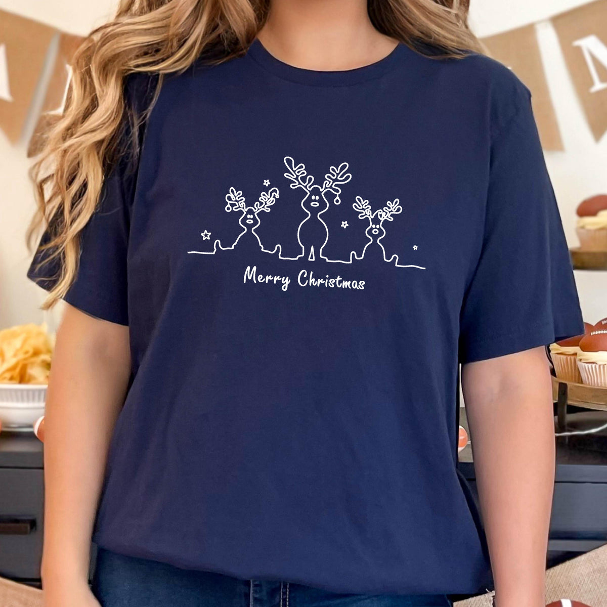 May include: Navy blue t-shirt featuring a white line drawing of three reindeer with star accents. The text "Merry Christmas" is written below the reindeer design. A festive holiday-themed apparel item.