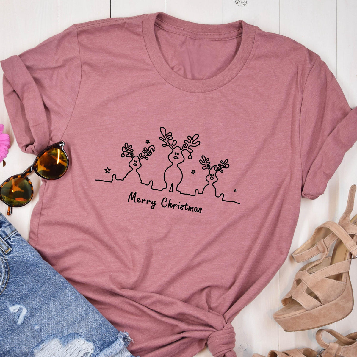 May include: A dusty rose t-shirt featuring a black line drawing of three reindeer with antlers and stars. The words "Merry Christmas" are written below the reindeer. The shirt is folded, with sunglasses and sandals visible.