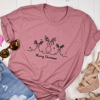 May include: A dusty rose t-shirt featuring a black line drawing of three reindeer with antlers and stars. The words "Merry Christmas" are written below the reindeer. The shirt is folded, with sunglasses and sandals visible.